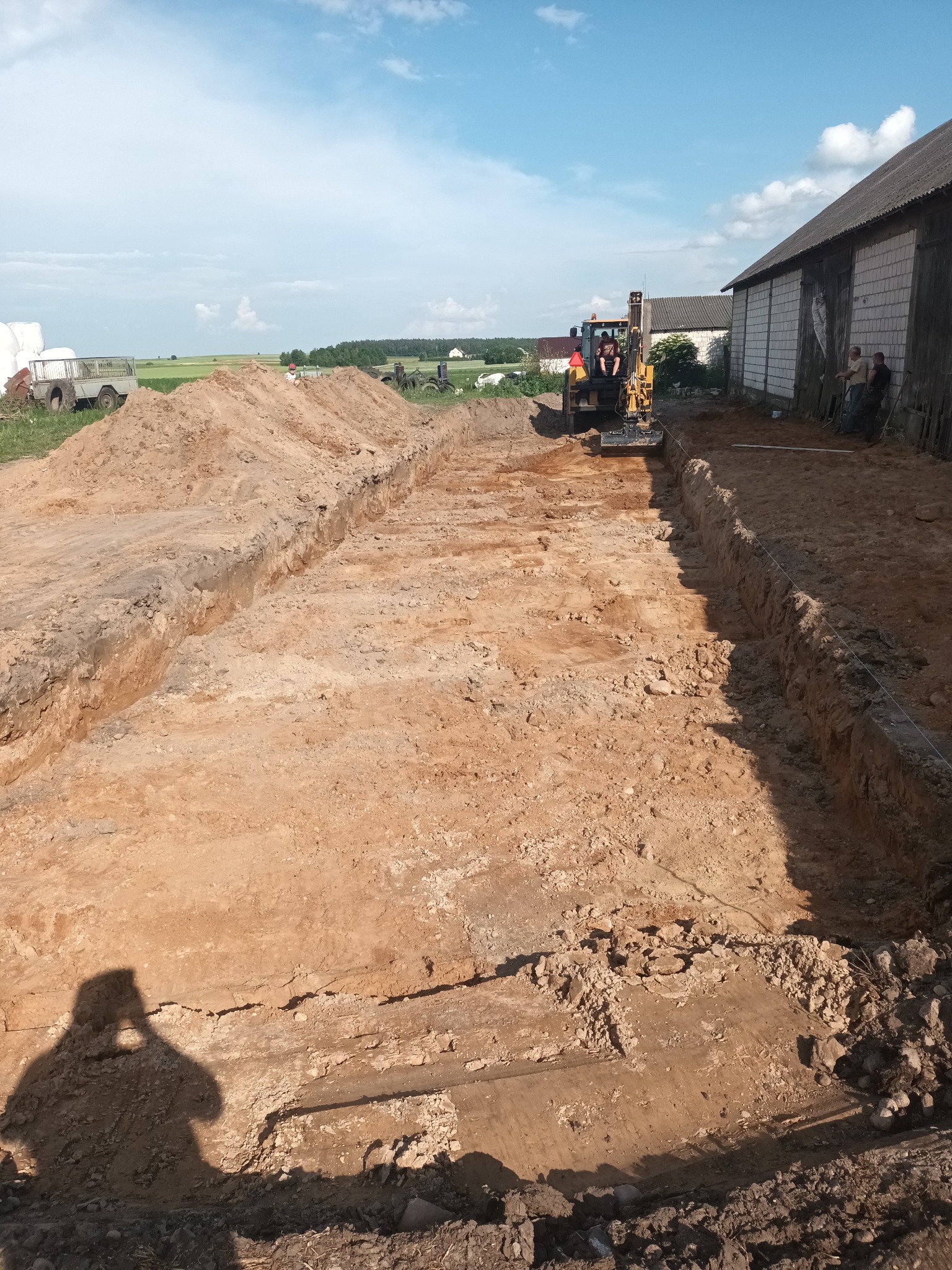 Żółta mini koparka w trakcie wykonywania wykopu liniowego w piaszczystej glebie, widoczny operator, w tle budynek gospodarczy i pole, cień fotografa w dolnej części kadru.