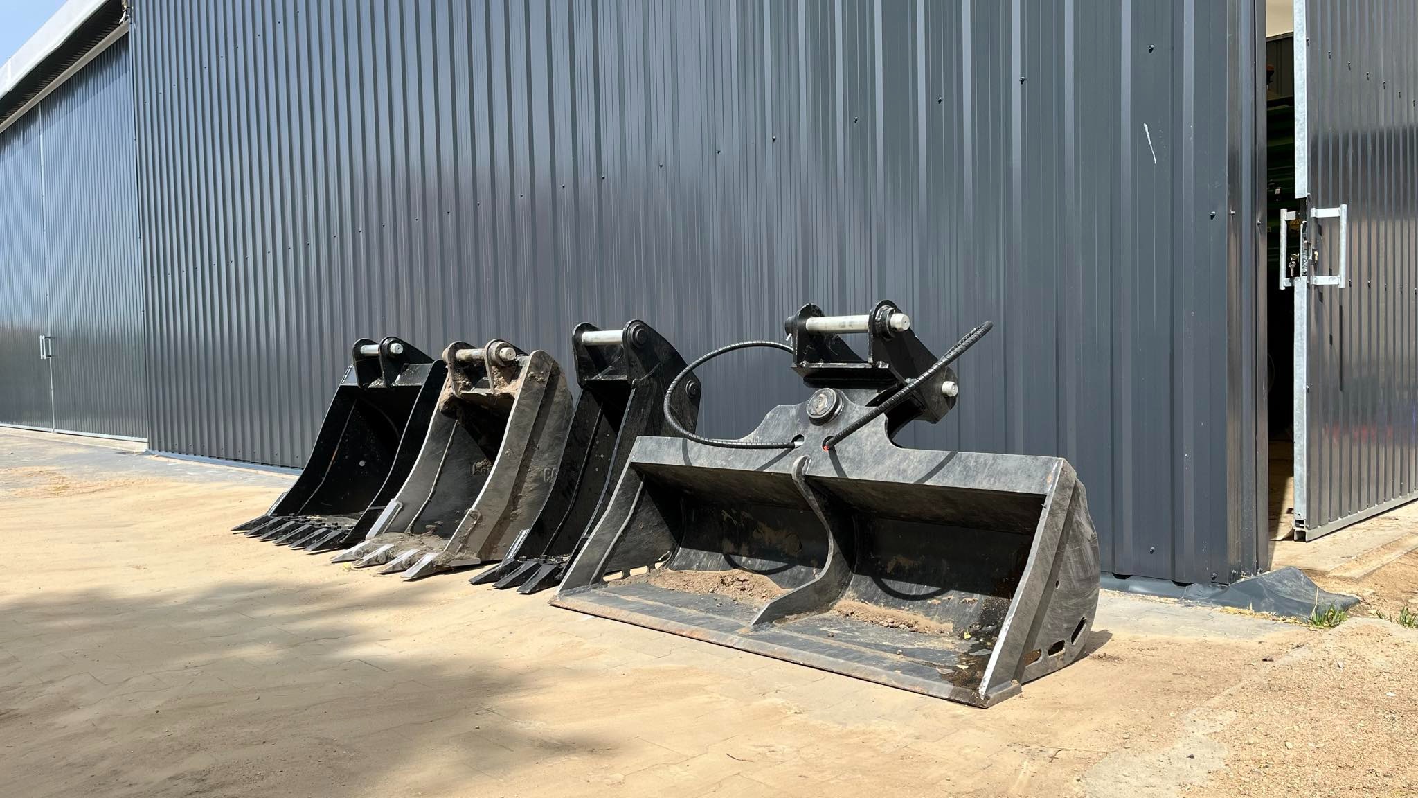 Rząd używanych, czarnych łyżek do koparko-ładowarki ustawionych przed szarą, metalową halą z otwartymi drzwiami.