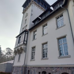 WS MONOLITH SPÓŁKA Z OGRANICZONĄ ODPOWIEDZIALNOŚCIĄ - Elewacja pałacu w Rogach po wymianie stolarki okiennej, widoczne nowe, białe ramy okienne z wieloma szybami w historycznym budynku z wieżą, kamienną podmurówką i elementami szachulcowymi.
