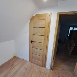 WS MONOLITH SPÓŁKA Z OGRANICZONĄ ODPOWIEDZIALNOŚCIĄ - Zamknięte, jasne drzwi wewnętrzne z poziomymi przeszkleniami i czarną klamką, zamontowane w skośnej ścianie poddasza, obok ościeżnica drugich drzwi z widocznym wnętrzem pokoju.