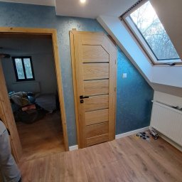 WS MONOLITH SPÓŁKA Z OGRANICZONĄ ODPOWIEDZIALNOŚCIĄ - Jasne, drewniane drzwi wewnętrzne z poziomymi panelami w nowym domu z oknem dachowym i niebieską ścianą z efektem dekoracyjnym, widoczne materiały montażowe na podłodze.