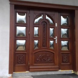 WS MONOLITH SPÓŁKA Z OGRANICZONĄ ODPOWIEDZIALNOŚCIĄ - Brązowe drzwi wejściowe z szybami i dekoracyjnymi elementami, zamontowane w surowym otoczeniu, z widocznymi śladami prac budowlanych na ścianie.