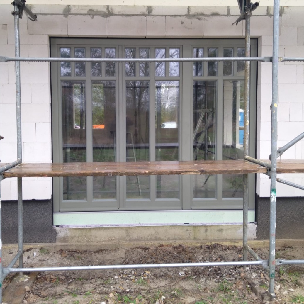 Montaż okna w surowym stanie, widoczne rusztowanie i bloczki betonowe, szare ramy okienne z podziałami.