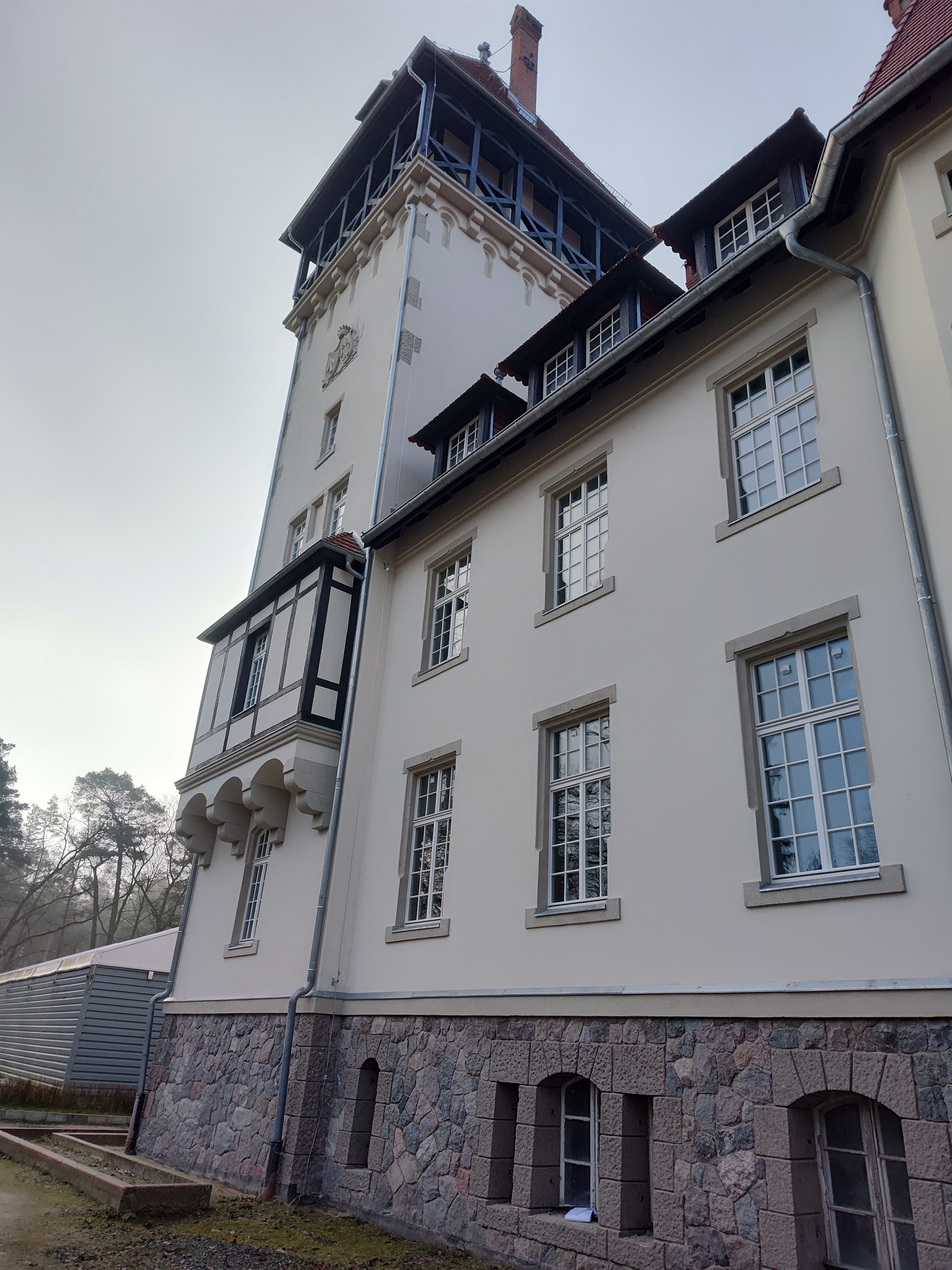 Elewacja pałacu w Rogach po wymianie stolarki okiennej, widoczne nowe, białe ramy okienne z wieloma szybami w historycznym budynku z wieżą, kamienną podmurówką i elementami szachulcowymi.