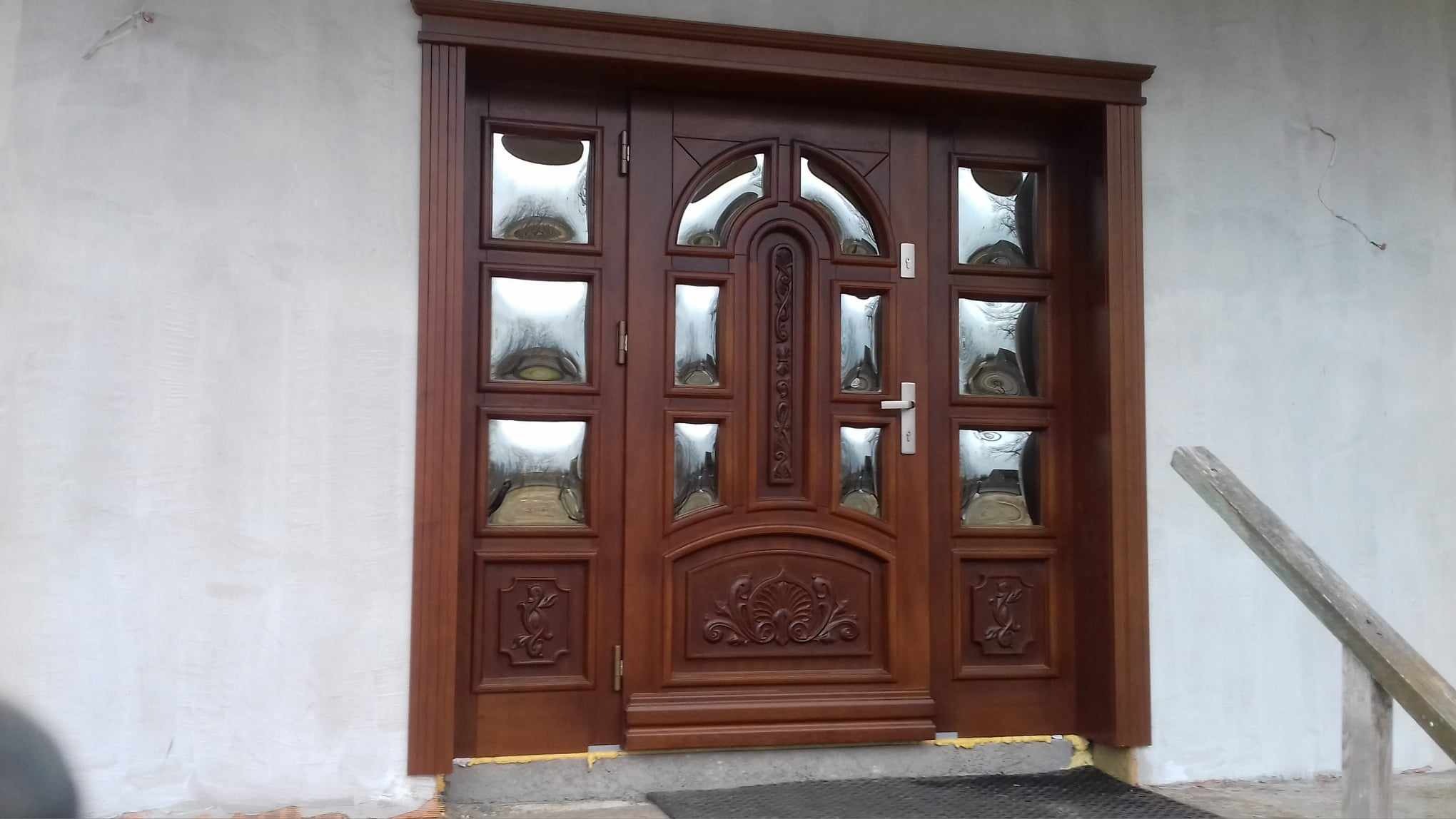 Brązowe drzwi wejściowe z szybami i dekoracyjnymi elementami, zamontowane w surowym otoczeniu, z widocznymi śladami prac budowlanych na ścianie.