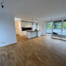 Dawid Papierz - Przestronne wnętrze po remoncie z parkietem ułożonym w jodełkę, białe ściany i wyspa kuchenna z czarnym blatem, widoczne niedokończone podłączenie lampy sufitowej.