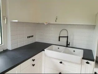 Biała kuchnia z czarnym blatem i baterią, białe płytki na ścianie ułożone w cegiełkę, dwukomorowy ceramiczny zlew farmerski, czarne uchwyty szafek.