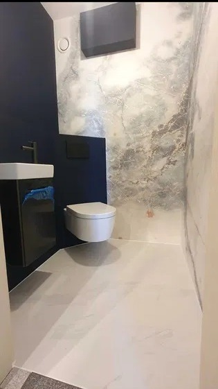 Nowoczesna łazienka z umywalką na szafce w kolorze ciemnego granatu, podwieszaną toaletą i ścianą wykończoną w imitacji marmuru.