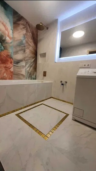 Nowoczesna łazienka z marmurową wanną obudowaną płytkami, dekoracyjną ścianą z motywem akwareli, podłogą z białych płyt z geometrycznym wzorem z złotych płytek i podświetlanym lustrem.