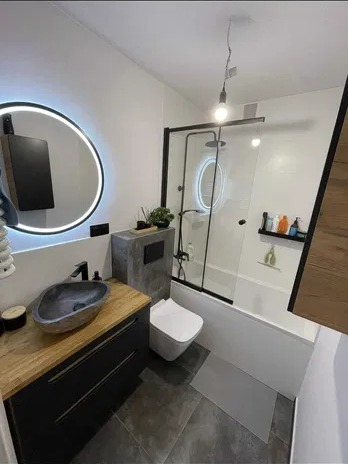 Nowoczesna łazienka z czarnymi akcentami: okrągłe lustro z podświetleniem LED, kamienna umywalka na drewnianym blacie, kabina prysznicowa z czarną ramą i wisząca lampa z żarówką Edisona.