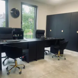 Nowoczesne biuro w czerni: biurka, krzesła z złotymi nogami, szafy i zegar na ścianie. Widok na zieleń za oknem. Minimalistyczny design, profesjonalne wnętrze.