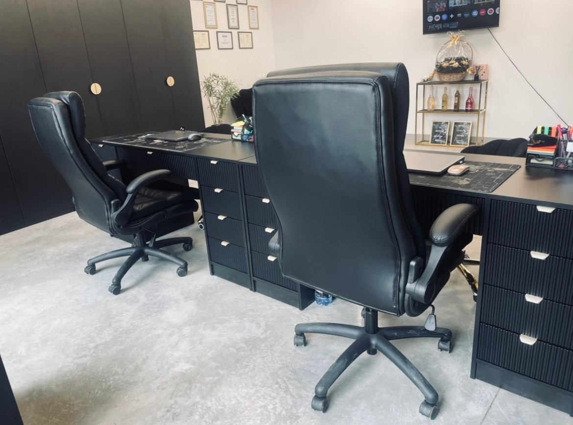 Nowoczesne biuro z czarnymi meblami: biurka z szufladami, skórzane fotele obrotowe. Na ścianie wiszą certyfikaty i dyplomy. W tle półka z dekoracjami.