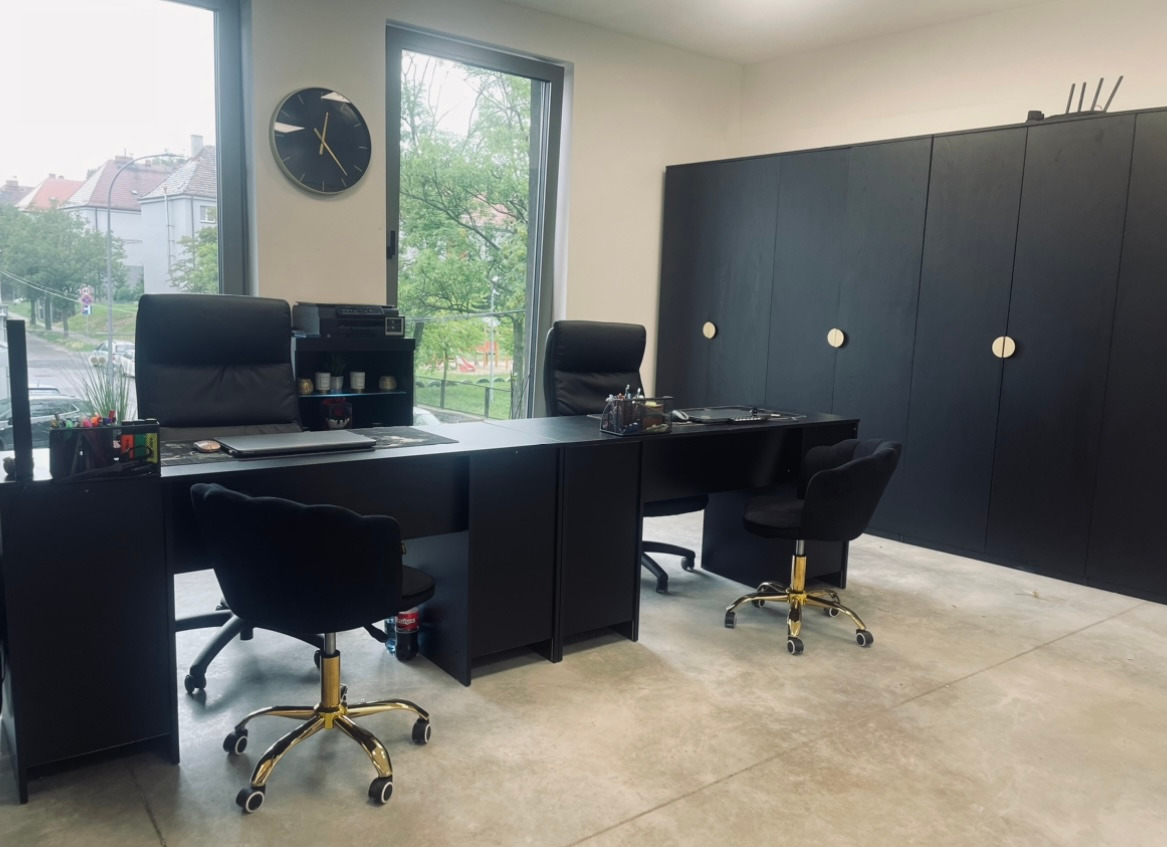 Nowoczesne biuro w czerni: biurka, krzesła z złotymi nogami, szafy i zegar na ścianie. Widok na zieleń za oknem. Minimalistyczny design, profesjonalne wnętrze.