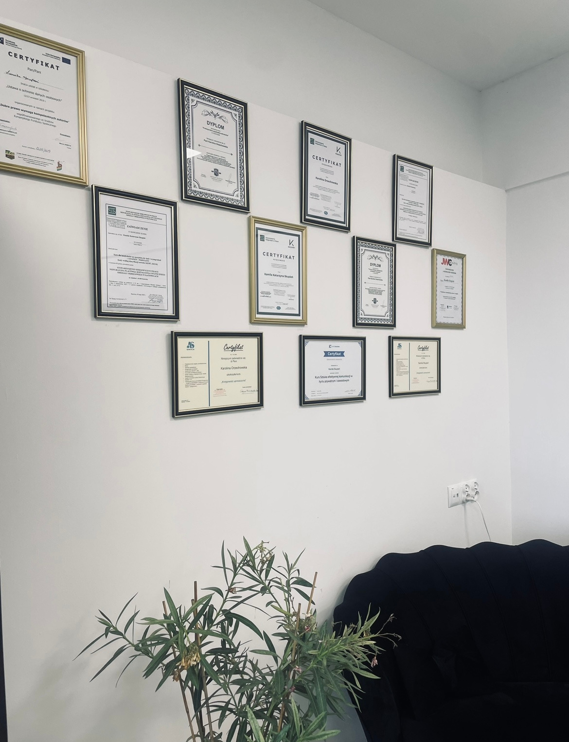 Ściana biura z ramkami zawierającymi certyfikaty i dyplomy, roślina doniczkowa i fragment czarnej sofy w dolnej części kadru. Prestiż i profesjonalizm.