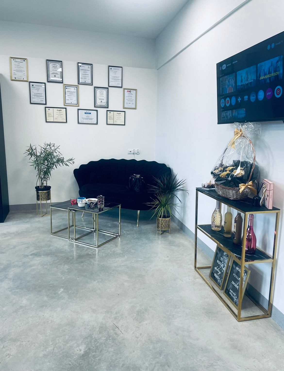 Eleganckie biuro z czarną sofą, złotymi stolikami i półką z dekoracjami. Na ścianie dyplomy i certyfikaty, a na półce tabliczki z motywacyjnymi hasłami.