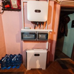 Mark-OZE sp z o. o. - Zamontowany biały inwerter Huawei z rozdzielnicą elektryczną w pomieszczeniu o różowych ścianach, z zapasem wody w niebieskich butelkach i gaśnicą w tle.