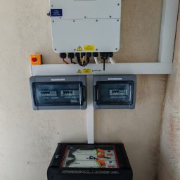 Mark-OZE sp z o. o. - Biały inwerter fotowoltaiczny marki Deye zamontowany na ścianie nad czarnym magazynem energii z widocznymi bateriami litowo-jonowymi oraz dwoma szarymi rozdzielnicami elektrycznymi.