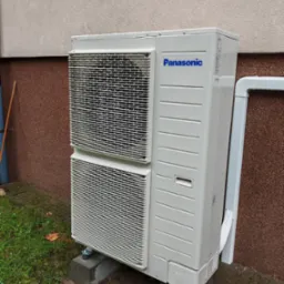 Zewnętrzna jednostka klimatyzacji Panasonic na betonowych podstawach, z widocznymi kroplami deszczu na obudowie i plastikową rurą odprowadzającą skropliny.
