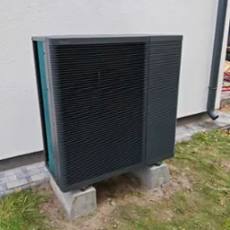 Zewnętrzna jednostka pompy ciepła Panasonic Aquarea, zamontowana na betonowych podstawach przy ścianie budynku z rynną spustową.