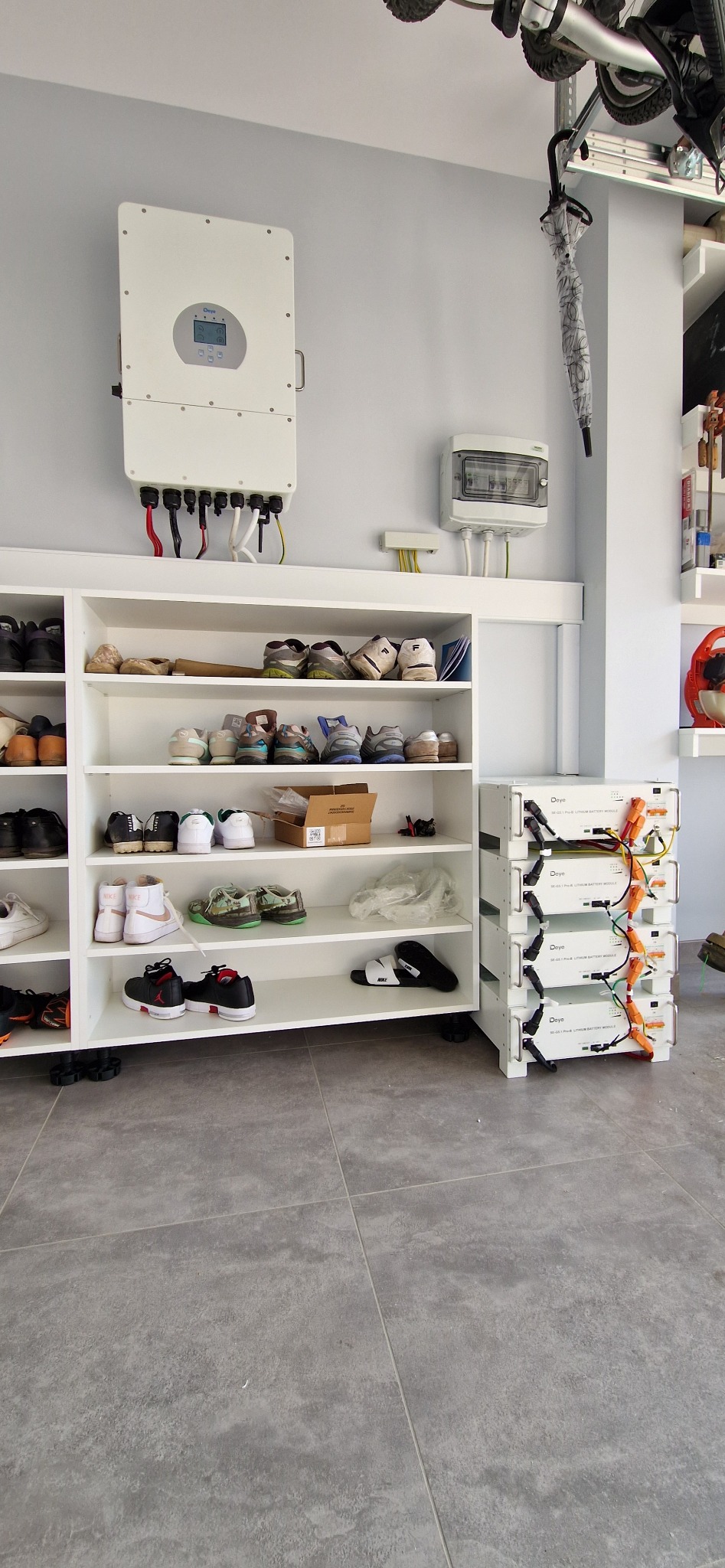 Biały inwerter fotowoltaiczny i moduły akumulatorowe Daye w garażu obok regału na buty, widoczne okablowanie i skrzynka elektryczna.
