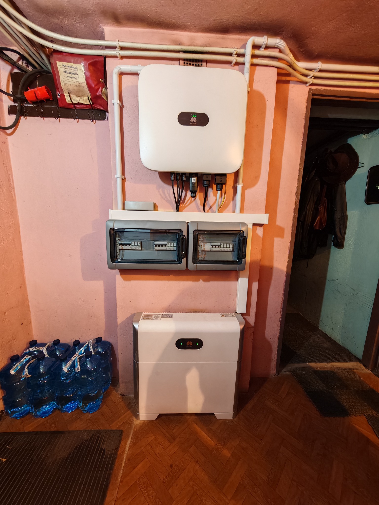 Zamontowany biały inwerter Huawei z rozdzielnicą elektryczną w pomieszczeniu o różowych ścianach, z zapasem wody w niebieskich butelkach i gaśnicą w tle.
