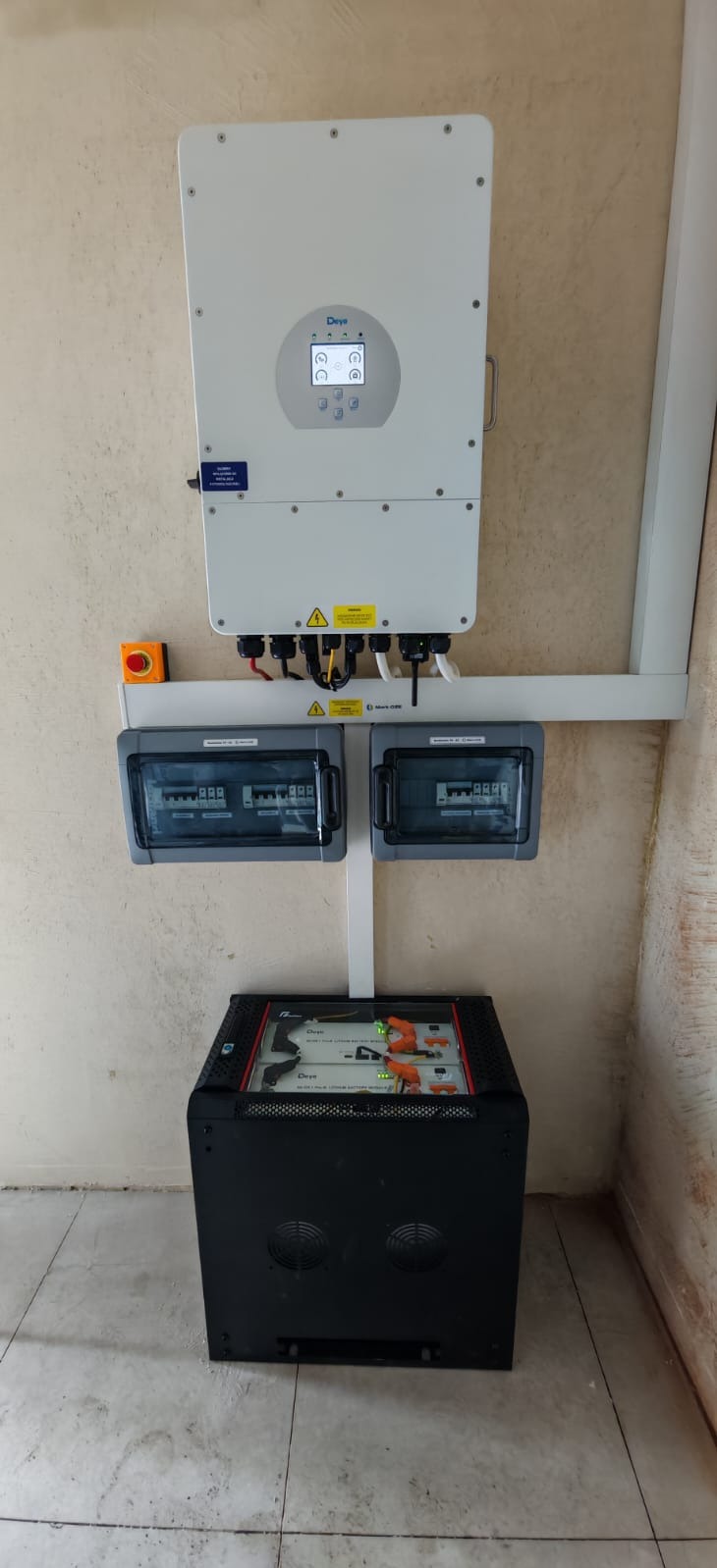 Biały inwerter fotowoltaiczny marki Deye zamontowany na ścianie nad czarnym magazynem energii z widocznymi bateriami litowo-jonowymi oraz dwoma szarymi rozdzielnicami elektrycznymi.