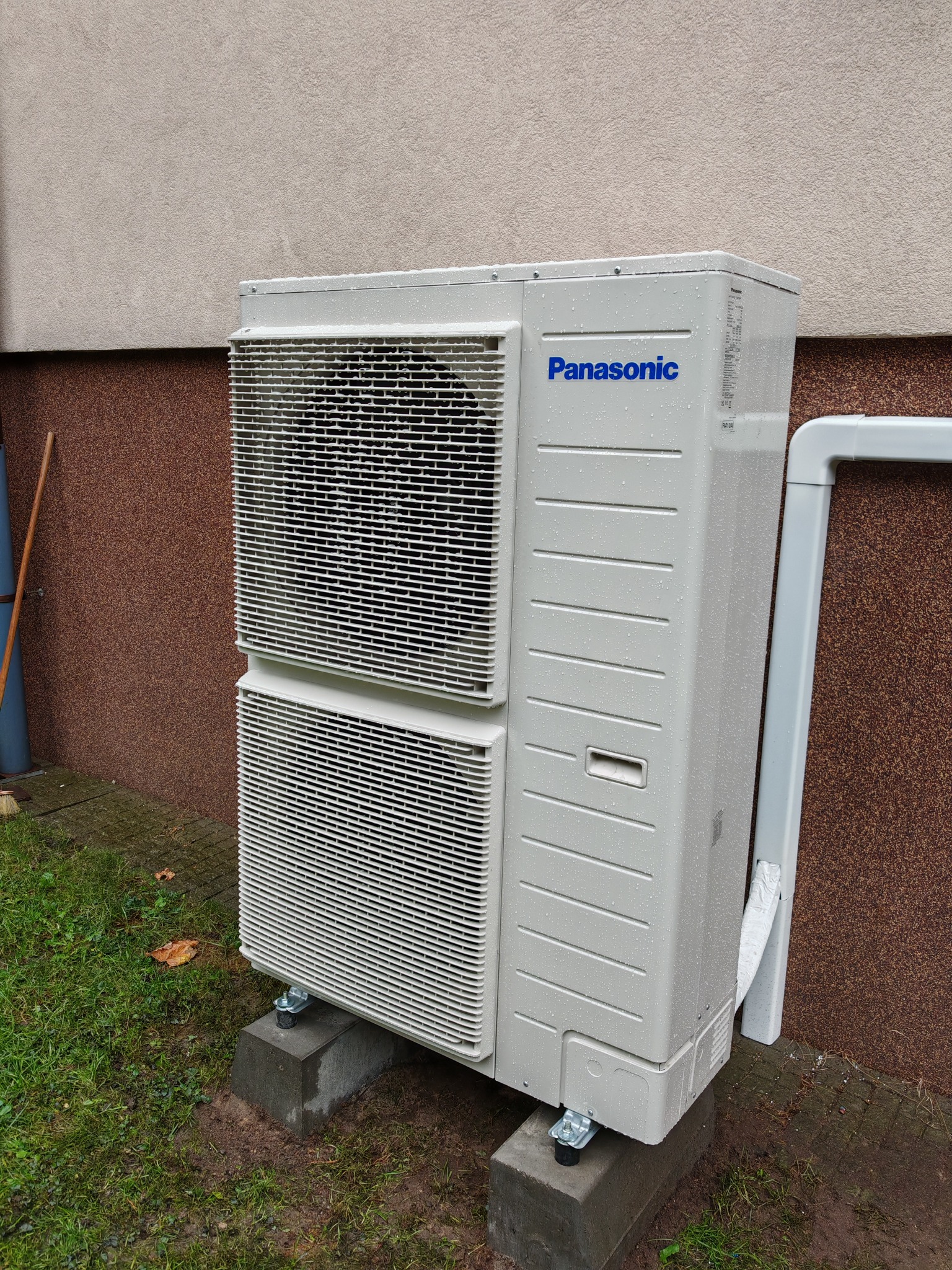Zewnętrzna jednostka klimatyzacji Panasonic na betonowych podstawach, z widocznymi kroplami deszczu na obudowie i plastikową rurą odprowadzającą skropliny.