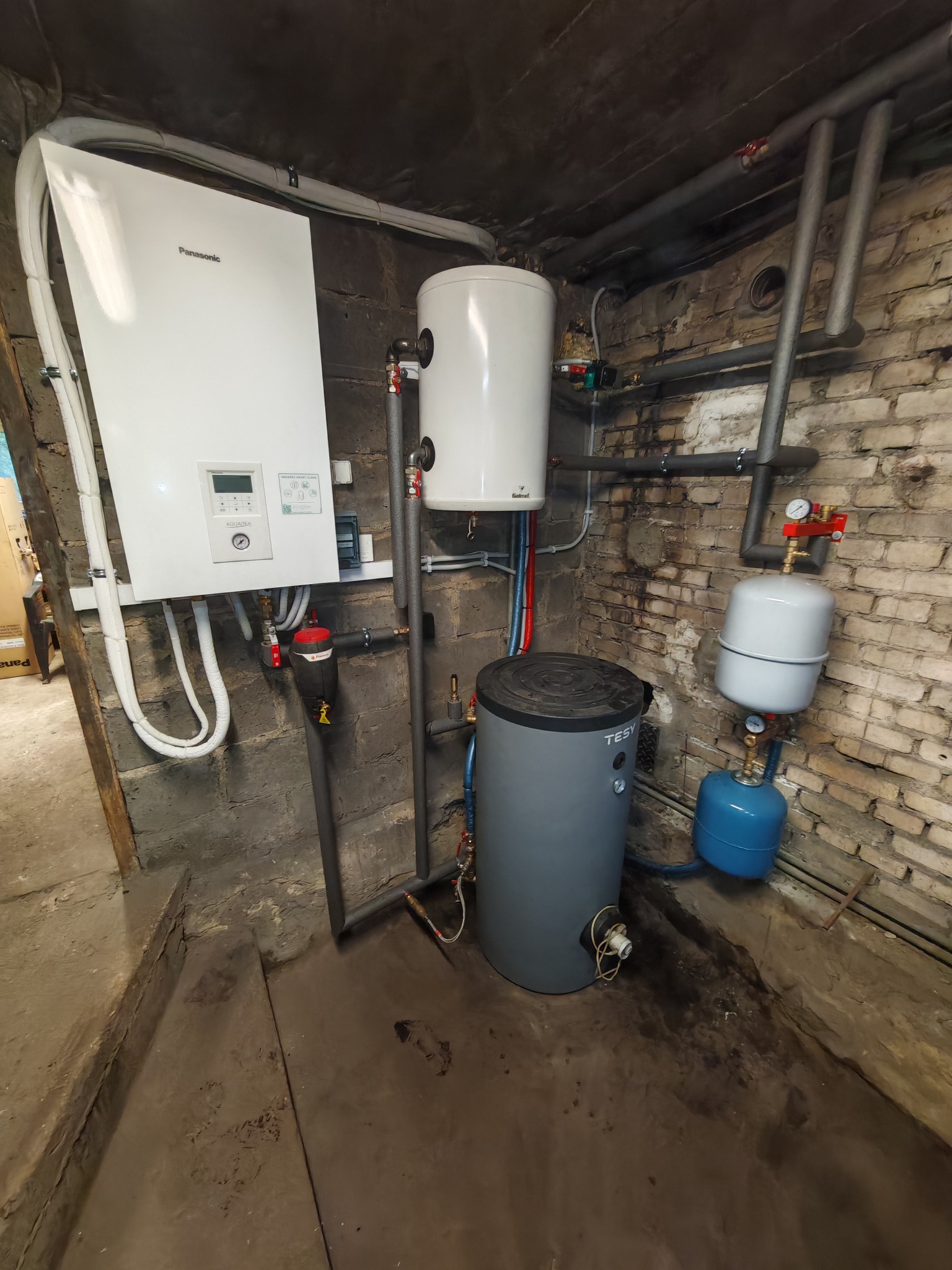 Zestaw urządzeń grzewczych w piwnicy: biała pompa ciepła Panasonic, zbiornik akumulacyjny Galmet, szary zasobnik TESY, oraz niebieski zbiornik wyrównawczy, widoczne rury instalacji grzewczej.