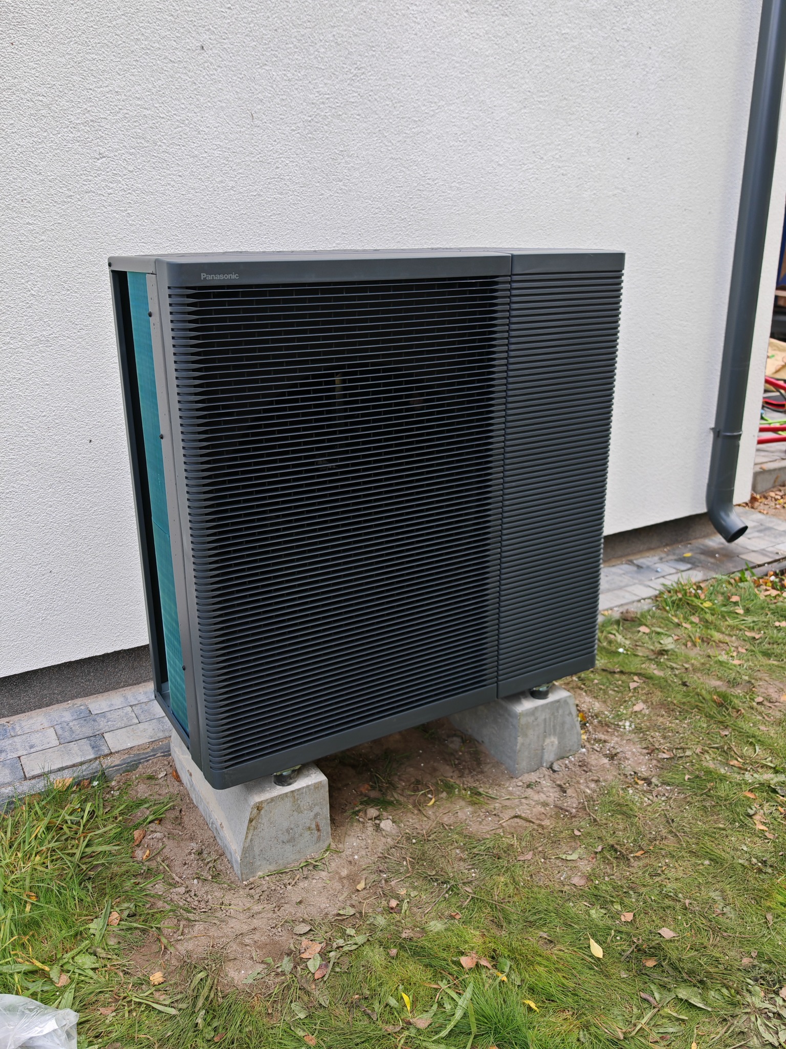 Zewnętrzna jednostka pompy ciepła Panasonic Aquarea, zamontowana na betonowych podstawach przy ścianie budynku z rynną spustową.