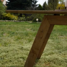 Stół z blatem z jasnego drewna i nogami w kształcie litery V, ustawiony na trawie w ogrodzie. Widok z dołu, podkreślający geometrię konstrukcji.