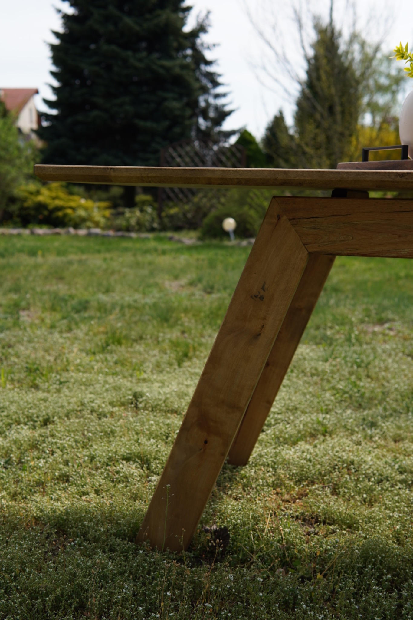 Stół z blatem z jasnego drewna i nogami w kształcie litery V, ustawiony na trawie w ogrodzie. Widok z dołu, podkreślający geometrię konstrukcji.
