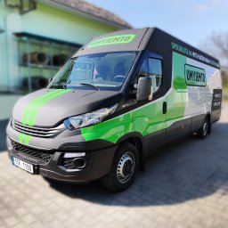 Agencja Promocyjno-Reklamowa SPOT s.c. Piotr Czerwiński, Wojciech Krawczyk - Czarno-szaro-zielony samochód dostawczy z logo firmy na tle budynku z półkami wypełnionymi przedmiotami.