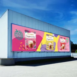 Billboard z trzema wariantami smakowymi Mokate Cappuccino na tle błękitnego nieba, umieszczony na ścianie budynku przemysłowego.