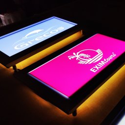 Agencja Promocyjno-Reklamowa SPOT s.c. Piotr Czerwiński, Wojciech Krawczyk - Dwa podświetlane panele reklamowe z logo firm: jeden z delfinem na niebieskim tle, drugi z palmą na różowym tle, oba na drewnianym blacie.