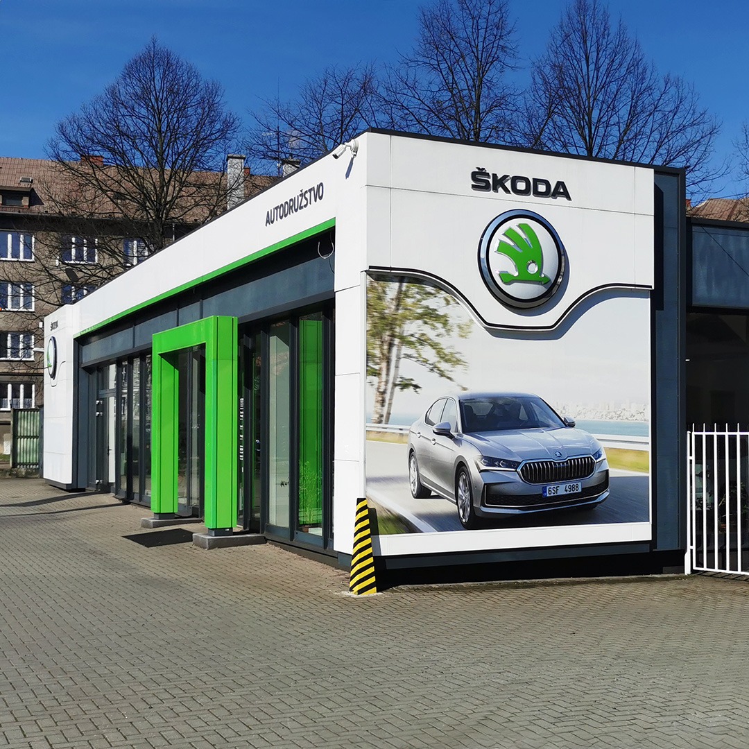 Oklejony budynek salonu samochodowego Skoda z dużym banerem reklamowym przedstawiającym srebrny samochód na tle krajobrazu.