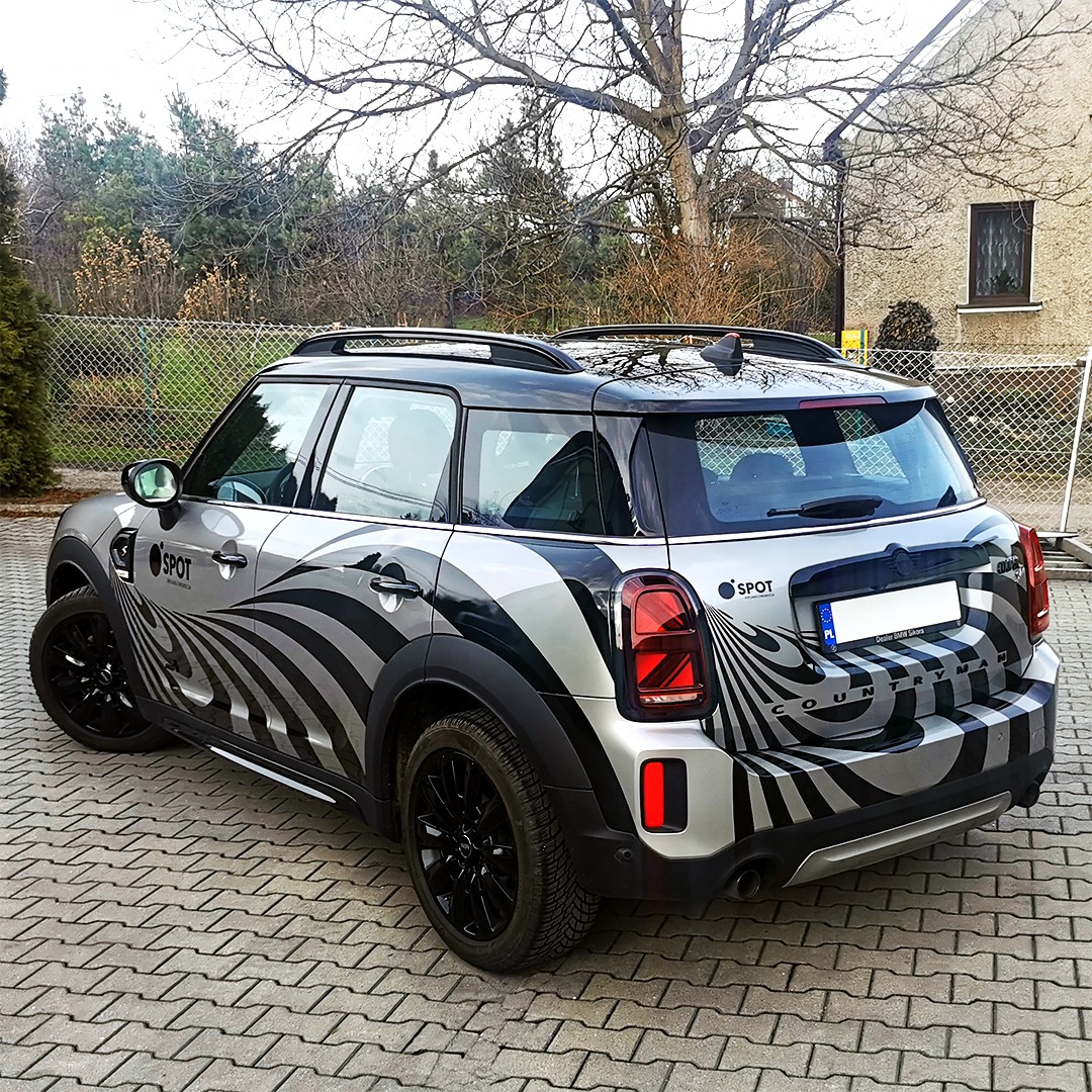 Srebrny Mini Countryman z czarnymi felgami i oklejeniem w czarno-srebrne, zakrzywione pasy tworzące iluzję optyczną, zaparkowany na kostce brukowej przed budynkiem.