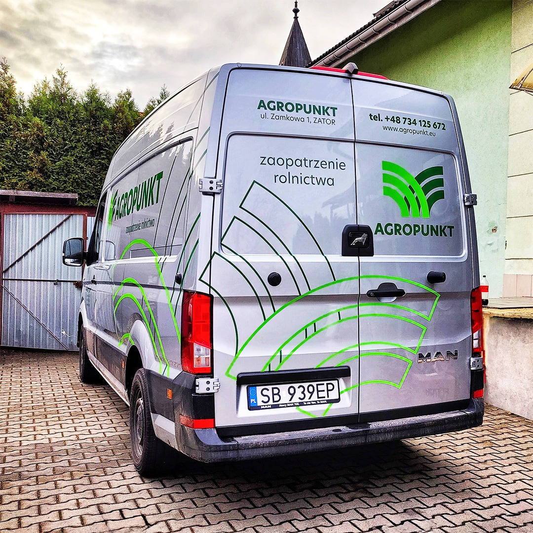 Srebrny samochód dostawczy marki MAN oklejony zieloną grafiką z logo firmy Agropunkt, zaparkowany na brukowanym podjeździe przed budynkiem.