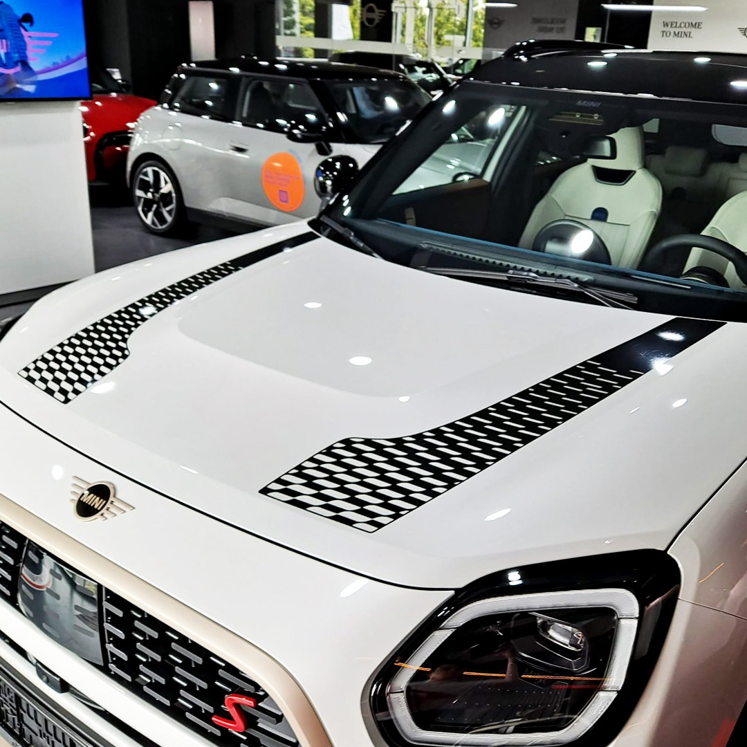 Biały samochód Mini Cooper z czarnymi paskami w szachownicę na masce, prezentowany w salonie samochodowym.