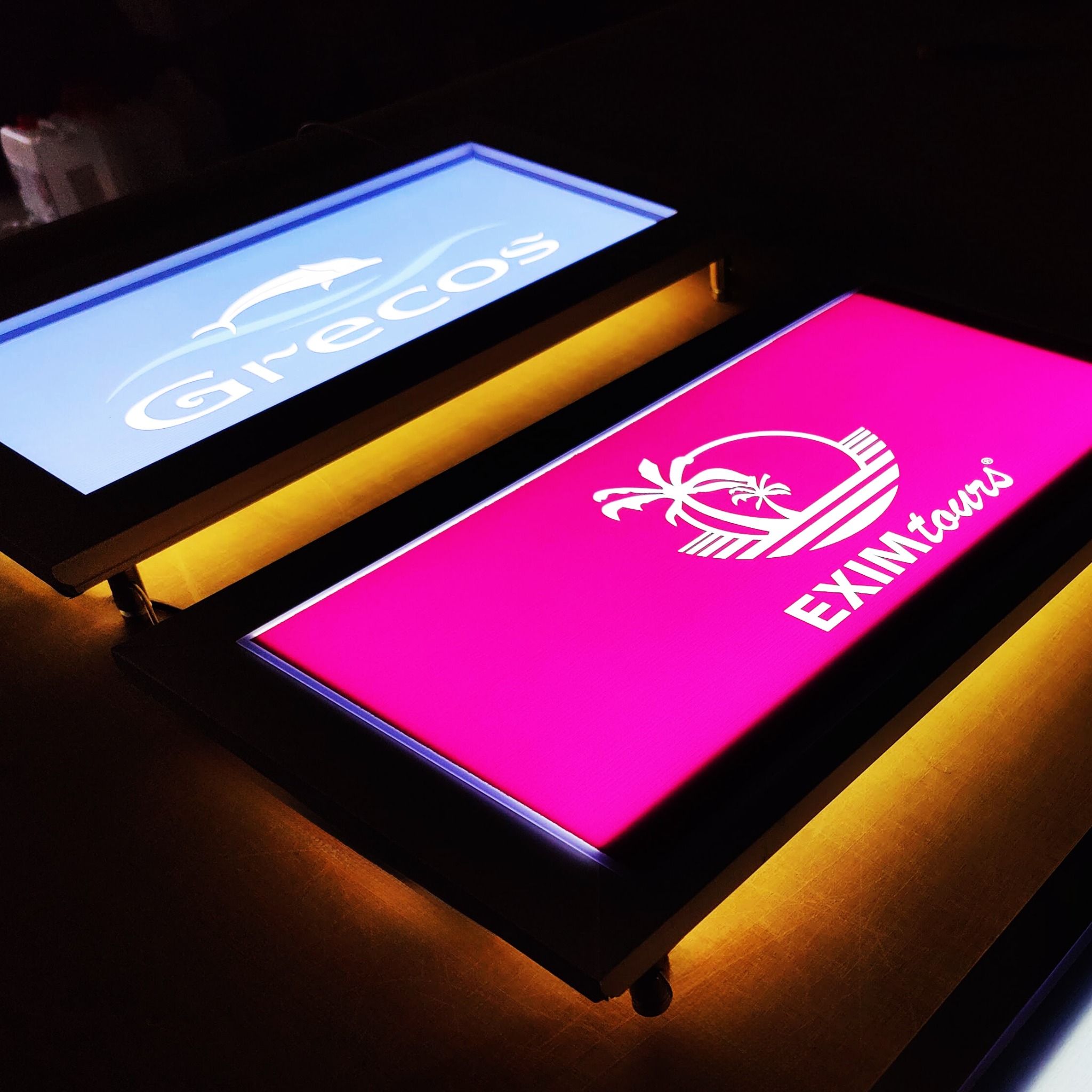 Dwa podświetlane panele reklamowe z logo firm: jeden z delfinem na niebieskim tle, drugi z palmą na różowym tle, oba na drewnianym blacie.