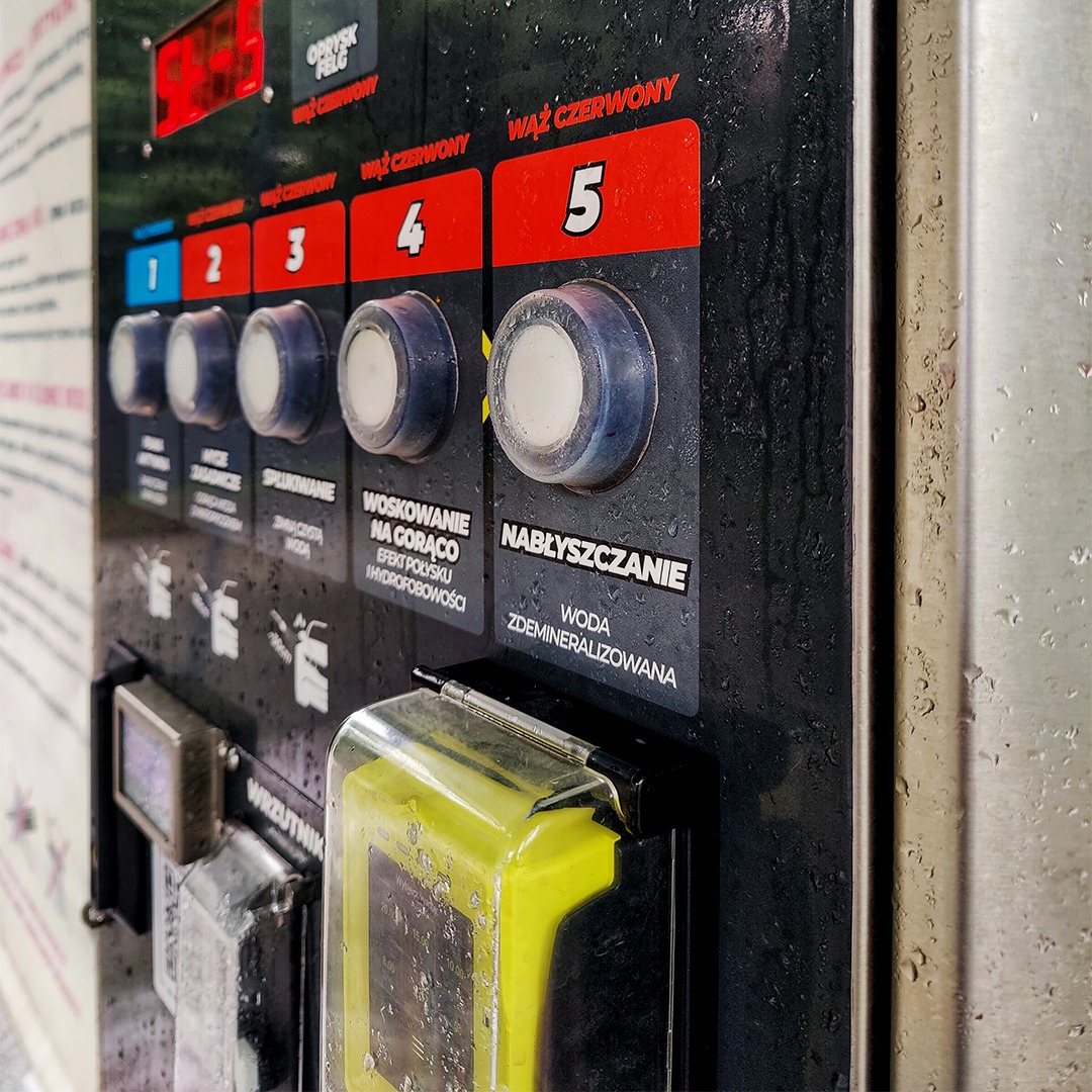 Panel sterowania myjni samochodowej z przyciskami wyboru programów: oprysk felg, wąż czerwony, spłukiwanie, woskowanie na gorąco, nabłyszczanie wodą zdemineralizowaną; widoczne krople wody.