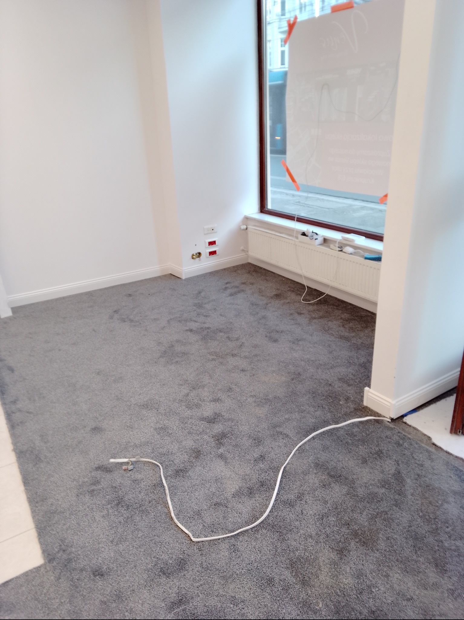 Pokój z szarą wykładziną dywanową, białymi ścianami, listwami przypodłogowymi i grzejnikiem pod oknem, widoczne gniazdka elektryczne i kabel na podłodze.
