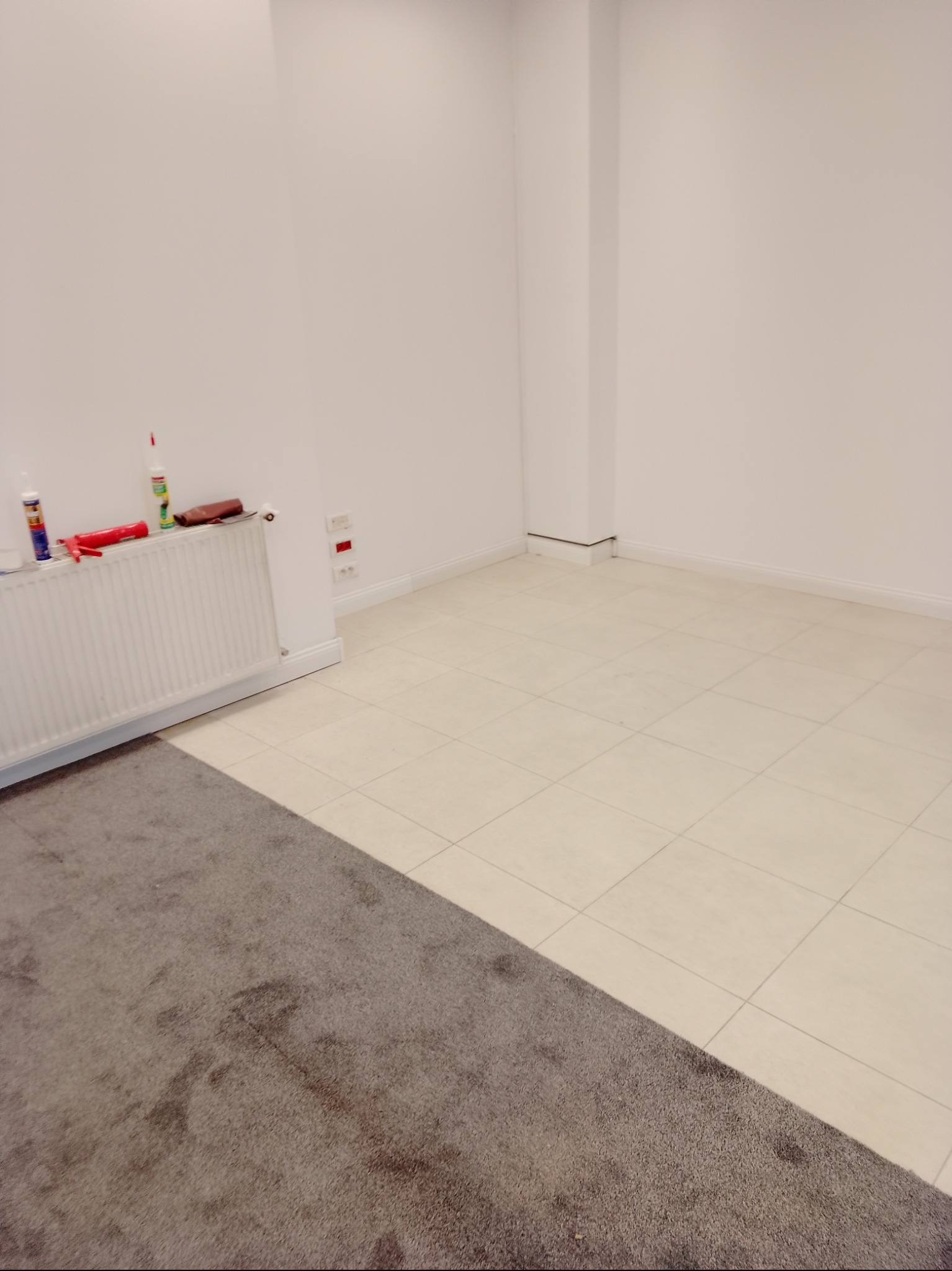 Wykończone wnętrze z białymi ścianami, jasnymi płytkami podłogowymi, szarym dywanem i grzejnikiem z akcesoriami do uszczelniania na parapecie.