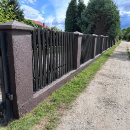Serwis Ogrodowy Wiśniewski Bartosz Wiśniewski - Długi, ciemny, drewniany płot z brązowymi, betonowymi słupkami i podstawą, oddzielający posesję od polnej drogi.
