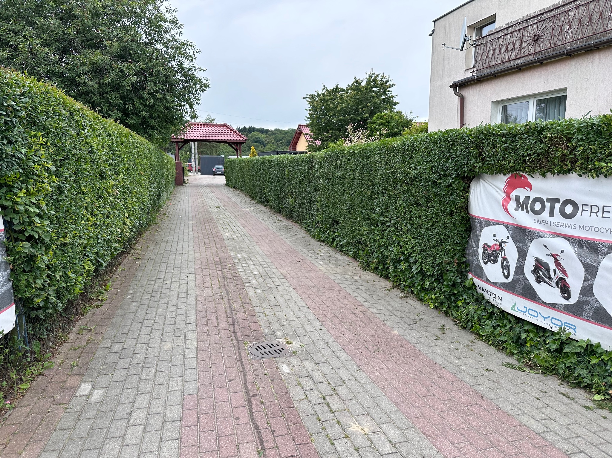 Długa aleja wyłożona kostką brukową prowadząca przez wysoki, równo przycięty żywopłot. Na żywopłocie widoczna reklama sklepu motocyklowego. W tle brama z czerwonym dachem i budynek mieszkalny.