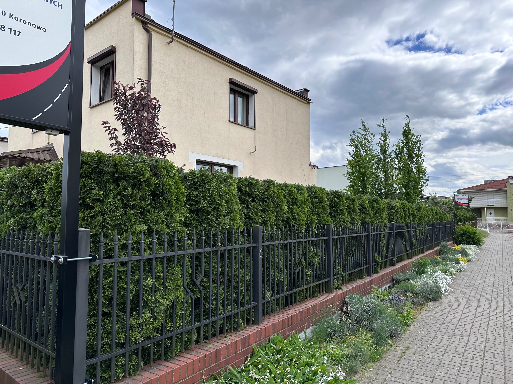 Równy szpaler gęstych, przystrzyżonych żywopłotów z tuj oddziela posesję od chodnika, za czarnym metalowym ogrodzeniem widać rabatę z kwitnącymi roślinami, w tle dwukondygnacyjny dom z beżową...