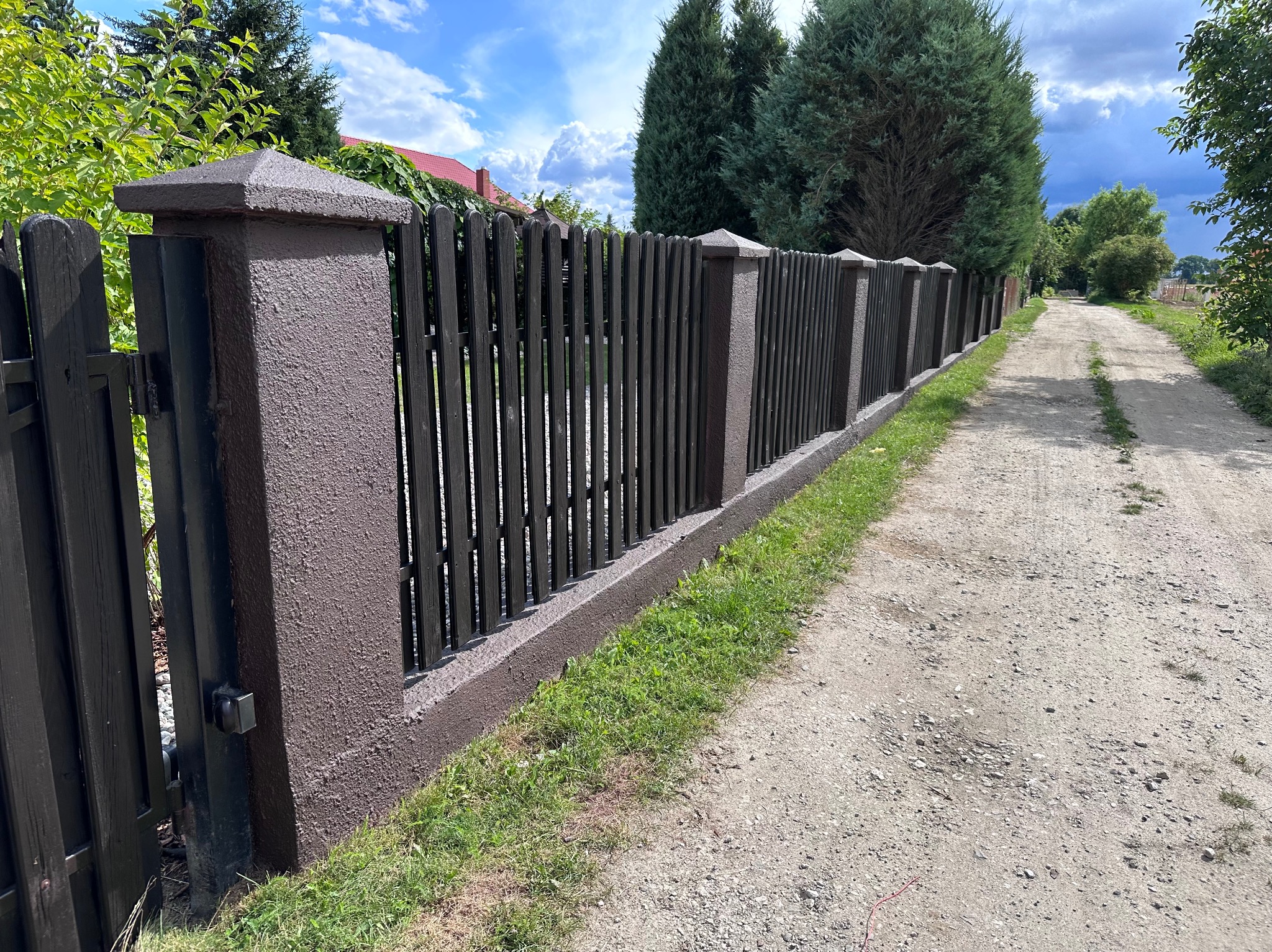 Długi, ciemny, drewniany płot z brązowymi, betonowymi słupkami i podstawą, oddzielający posesję od polnej drogi.