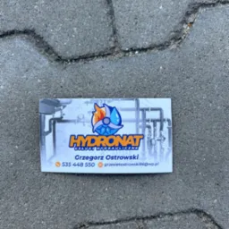 Wizytówka firmy hydraulicznej Hydronat leżąca na szarej kostce brukowej, z logo przedstawiającym płomień i kroplę wody, numerem telefonu i adresem e-mail Grzegorza Ostrowskiego.