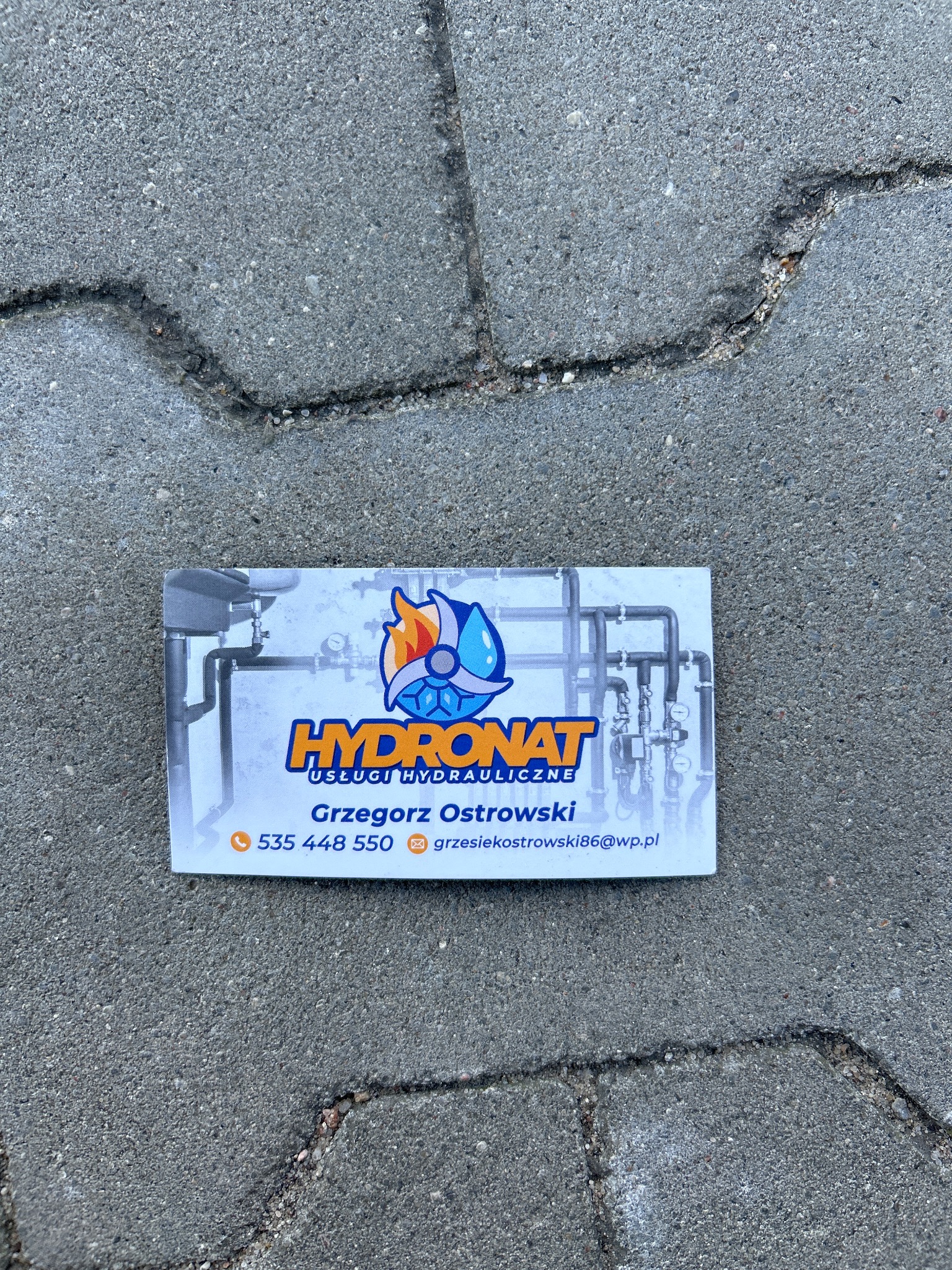Wizytówka firmy hydraulicznej Hydronat leżąca na szarej kostce brukowej, z logo przedstawiającym płomień i kroplę wody, numerem telefonu i adresem e-mail Grzegorza Ostrowskiego.