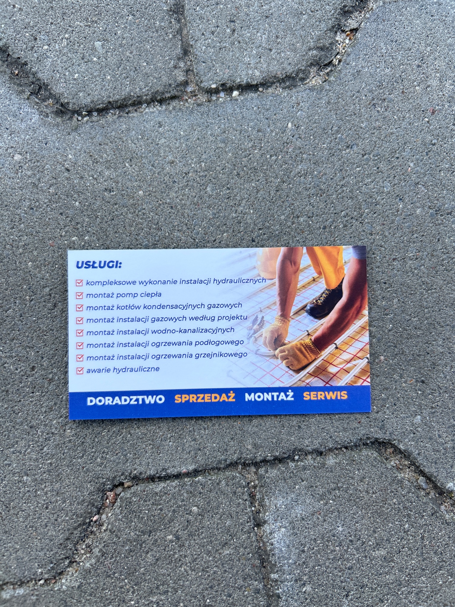 Wizytówka firmy instalacyjnej leżąca na brukowanej nawierzchni, prezentująca ofertę usług hydraulicznych i grzewczych, w tle fragment zdjęcia z montażu ogrzewania podłogowego.