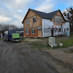 Dom w trakcie ocieplania, widoczne rusztowania, częściowo ocieplona ściana, zaparkowany van z napisem 'Izolacje natryskowe pianą', obok przyczepka z agregatem natryskowym.